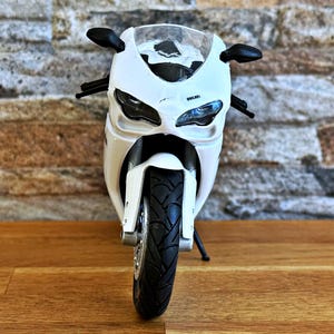 K&ouml;nnte beinhalten: Wei&szlig;es Ducati Motorradmodell mit schwarzen Akzenten, einschlie&szlig;lich Spiegeln und Reifen. Das Modell tr&auml;gt den Schriftzug "Ducati" auf der Vorderseite. Das Motorrad steht auf einer Holzoberfl&auml;che.