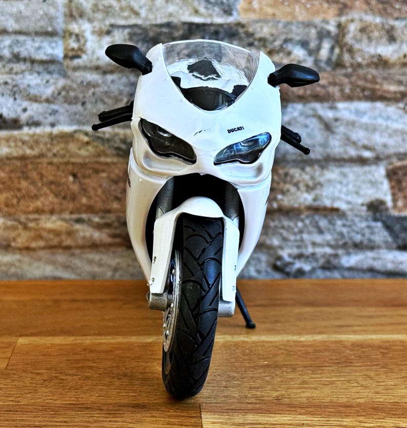 K&ouml;nnte beinhalten: Wei&szlig;es Ducati Motorradmodell mit schwarzen Akzenten, einschlie&szlig;lich Spiegeln und Reifen. Das Modell tr&auml;gt den Schriftzug "Ducati" auf der Vorderseite. Das Motorrad steht auf einer Holzoberfl&auml;che.