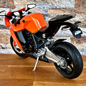 May include: Orange and black motorcycle model with a black seat and a silver exhaust pipe. The motorcycle has a black and white KTM logo on the back.