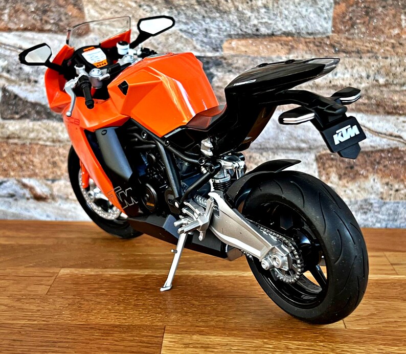 May include: Orange and black motorcycle model with a black seat and a silver exhaust pipe. The motorcycle has a black and white KTM logo on the back.