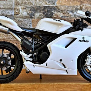 K&ouml;nnte beinhalten: Wei&szlig;es Ducati 1198 Motorradmodell mit schwarzen R&auml;dern und Akzenten. Das Motorrad hat das Ducati-Logo auf dem Tank und die Nummer 1198 auf der Seitenverkleidung. Das Modell wird auf einer Holzoberfl&auml;che pr&auml;sentiert.