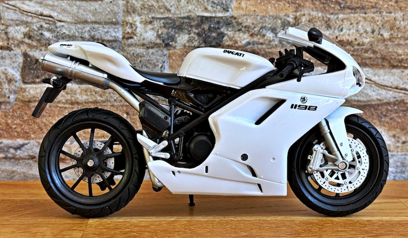 K&ouml;nnte beinhalten: Wei&szlig;es Ducati 1198 Motorradmodell mit schwarzen R&auml;dern und Akzenten. Das Motorrad hat das Ducati-Logo auf dem Tank und die Nummer 1198 auf der Seitenverkleidung. Das Modell wird auf einer Holzoberfl&auml;che pr&auml;sentiert.