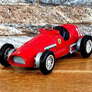 May include: A red vintage race car model with the number 10 on the side. The car has black tires, silver rims, and a Ferrari logo. The model is on a wooden surface.