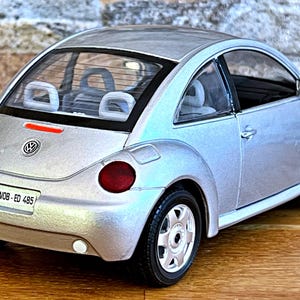Könnte beinhalten: Silbernes Modell eines Volkswagen Käfers mit roten Rücklichtern und einem Nummernschild mit der Aufschrift "WOB-ED 485". Das Spielzeugauto hat eine detaillierte Innenausstattung und steht auf einer Holzoberfläche.