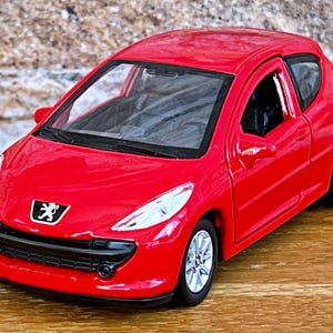 Puede incluir: Un coche de juguete rojo brillante con ruedas plateadas y una parrilla negra. El coche tiene el logotipo de Peugeot y está sobre una superficie de madera. La carrocería es de color rojo vivo.