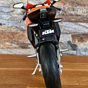 May include: A black and orange KTM motorcycle model with a black rear fender and a black rear tire. The motorcycle is on a wooden surface.