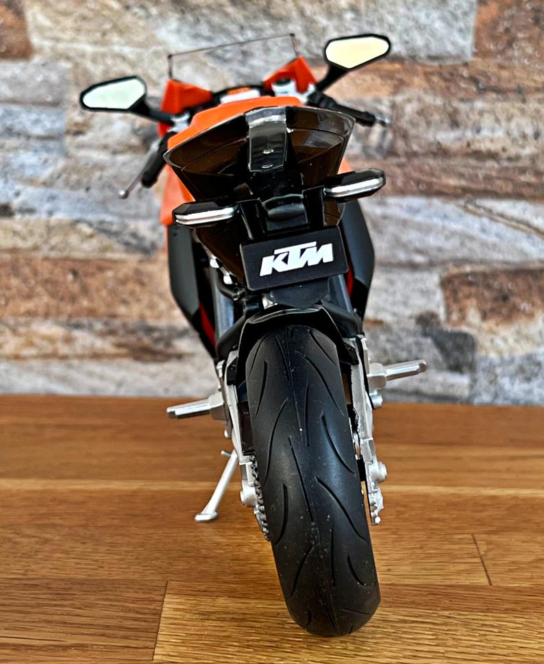 May include: A black and orange KTM motorcycle model with a black rear fender and a black rear tire. The motorcycle is on a wooden surface.