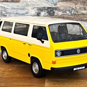 May include: A yellow and white Volkswagen Type 2 (T3) van model. The van has black tires, a black front grill, and a black bumper. The model is sitting on a wooden surface with a brick wall in the background.