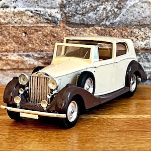 Könnte beinhalten: Ein cremefarbenes und braunes Oldtimer-Modell mit detailliertem Frontgrill, Scheinwerfern und Kotflügeln. Das klassische Auto steht auf einer Holzoberfläche vor einer Backsteinmauer. Das Modellauto ist ein Sammlerstück.