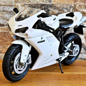 Peut inclure: Modèle réduit de moto Ducati 1198 blanche avec des pneus et des détails noirs. La moto a un pare-brise transparent et le logo Ducati est visible. Le modèle est posé sur une surface en bois avec un fond en briques.