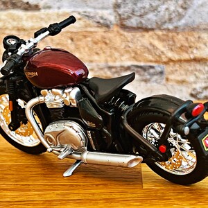 May include: A red and black Triumph motorcycle model with chrome accents. The motorcycle has a single seat and a black rear fender with a red taillight.