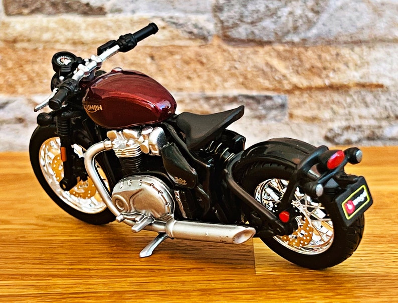 May include: A red and black Triumph motorcycle model with chrome accents. The motorcycle has a single seat and a black rear fender with a red taillight.