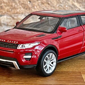 Puede incluir: Un modelo de coche de juguete rojo de un Range Rover Evoque. El coche tiene una parrilla negra y ruedas negras con detalles plateados.