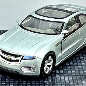 May include: A silver toy car model of a Chevrolet Volt concept car. The car has a black interior and a clear plastic roof. The car is sitting on a black and white checkered surface.