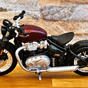 May include: A burgundy and black Triumph motorcycle model with chrome accents. The motorcycle has a black seat and a chrome exhaust pipe.