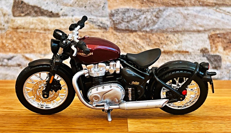 May include: A burgundy and black Triumph motorcycle model with chrome accents. The motorcycle has a black seat and a chrome exhaust pipe.