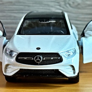May include: A white toy car with a black roof and open doors. The car has a black grill with the Mercedes-Benz logo. The car is on a wooden surface.