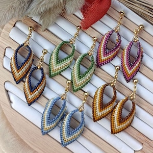 Può includere: Sei paia di orecchini con perline di vari colori, tra cui blu, verde, marrone e viola. Gli orecchini sono esposti su un supporto in legno con sfondo bianco.