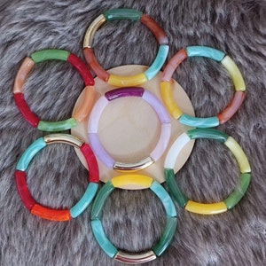 Peut inclure: Un ensemble de huit bracelets colorés en verre rond, plat et texturé. Les bracelets sont disposés en cercle sur une surface brun clair. Chaque bracelet est doté d'une bande métallique dorée.