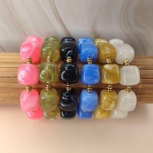 Peut inclure: Six bracelets disposés côte à côte, chacun composé de perles carrées en résine marbrée rose, vert olive, noir, bleu, doré et blanc. De petites perles intercalaires rondes de couleur or métallique sont placées entre chaque perle en résine.