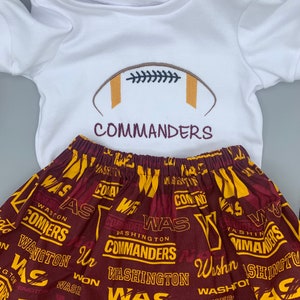 May include: A white onesie with a brown football and the text "COMMANDERS" embroidered on the front. A burgundy skirt with a repeating pattern of the text "WASHINGTON COMMANDERS" in yellow and burgundy.