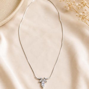 May include: A silver-toned necklace featuring a delicate chain and a central pendant composed of three clear, marquise-cut stones arranged in a leaf-like cluster, secured with a small metal clasp at the back of the neck.