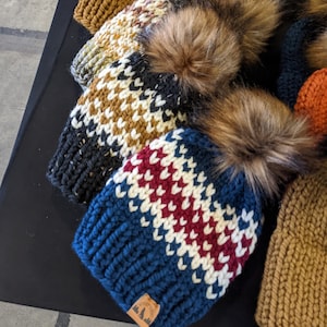 以下が含まれることがあります： 黒、白、茶色、緑、赤、青など、さまざまな色の組み合わせで、フェイクファーのポンポンが付いたニット帽が5つあります。帽子は暗い表面に一列に並んでいます。