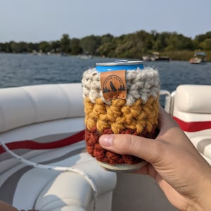May include: A crocheted beverage holder with a white, yellow, and red color scheme. The holder has a brown leather patch with the text "Nature Nomad" and a wolf silhouette.