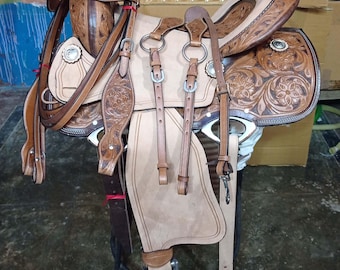 May include: A brown leather saddle with intricate floral tooling and a light brown suede seat. The saddle is complete with stirrups, a cinch, and a breast collar.