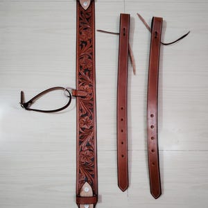 Puede incluir: Un collar de pecho de cuero marrón para caballo con estampado floral. El collar de pecho tiene dos correas ajustables con hebillas.