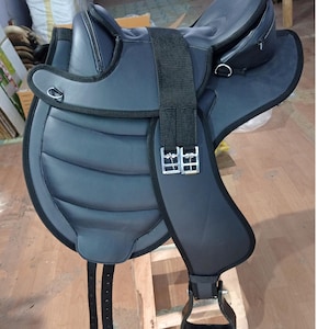 Puede incluir: Una silla de montar inglesa negra con asiento acolchado y rodilleras. La silla de montar presenta una cincha negra con hebillas plateadas y estribos. La silla de montar está diseñada para la comodidad y el rendimiento.