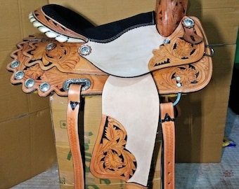 May include: A brown leather saddle with intricate floral carving and a black suede seat. The saddle has silver accents and stirrups.