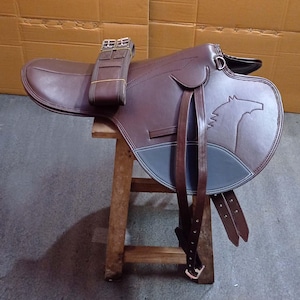 Op de afbeelding: Een bruin leren paardenzadel met een paardenhoofd ontwerp. Het zadel heeft een donkerblauwe accentkleur en bruine riemen. Het staat op een kleine houten kruk.