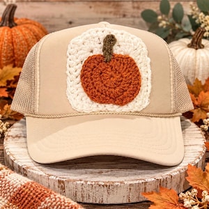 Puede incluir: Una gorra tipo camionero de color beige con un parche cuadrado de ganchillo color crema en el panel frontal. El parche muestra una calabaza de ganchillo texturizada, redonda y de color naranja quemado con un pequeño tallo verde oscuro.
