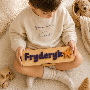 Puede incluir: Un ni&ntilde;o con una sudadera y pantalones cortos beige sostiene un rompecabezas de nombre de madera. El rompecabezas deletrea "Fryderyk" en letras azules y presenta una estrella de mar naranja. Tambi&eacute;n se ve un coche de juguete de madera y un oso de peluche.