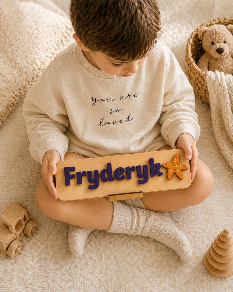Puede incluir: Un ni&ntilde;o con una sudadera y pantalones cortos beige sostiene un rompecabezas de nombre de madera. El rompecabezas deletrea "Fryderyk" en letras azules y presenta una estrella de mar naranja. Tambi&eacute;n se ve un coche de juguete de madera y un oso de peluche.