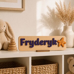 Puede incluir: Un letrero de nombre de madera con el nombre "Fryderyk" en letras azul oscuro y una estrella de mar naranja. El letrero est&aacute; sobre una estanter&iacute;a blanca con un conejito de peluche, un juguete de apilamiento de madera y un jarr&oacute;n con flores secas.