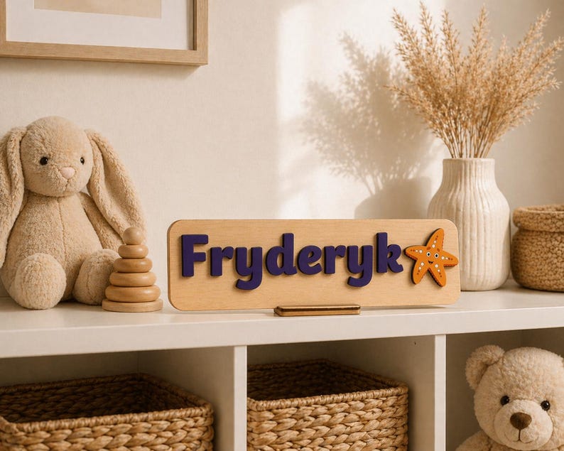 Puede incluir: Un letrero de nombre de madera con el nombre "Fryderyk" en letras azul oscuro y una estrella de mar naranja. El letrero est&aacute; sobre una estanter&iacute;a blanca con un conejito de peluche, un juguete de apilamiento de madera y un jarr&oacute;n con flores secas.