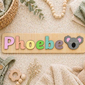 Puede incluir: Rompecabezas de madera con el nombre "Phoebe" en letras coloridas y un koala. El rompecabezas est&aacute; rodeado de ropa de beb&eacute;, un conejito de peluche y juguetes de madera sobre una alfombra crema.