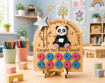 Puede incluir: Un cuadro de recompensas de madera con una ilustración de panda y el texto "Sophie's Reward Chart Caught You Being Good!". El cuadro tiene fichas en forma de estrella en rosa y azul. También presenta una estrella plateada y un diseño de bambú.