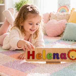 Puede incluir: Un rompecabezas de madera con el nombre "Helena" en letras coloridas. El rompecabezas incluye un gr&aacute;fico de le&oacute;n y se muestra sobre una alfombra colorida. La ni&ntilde;a lleva un su&eacute;ter blanco y pantalones rosas.