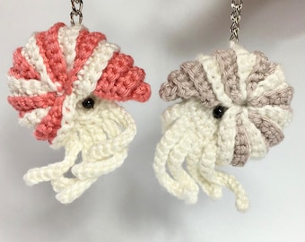 May include: Two handmade crochet nautilus shell keychains. One features a pink and white striped shell, the other a gray and white striped shell. Both have cream-colored tentacles and black bead eyes, attached to silver-tone chains.