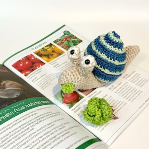May include: A handmade crochet snail with a blue and green striped shell and beige body rests on a gardening magazine. A small green crocheted lettuce is also present. The magazine displays tomato varieties.