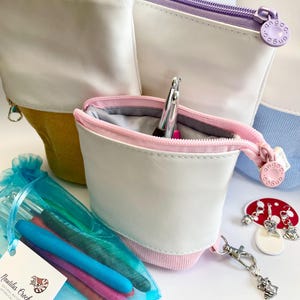 May include: Various crochet accessories are displayed, including white, yellow, and purple zippered pouches. A blue mesh bag holds crochet hooks in different colours. A mushroom charm and a frog charm are also present.