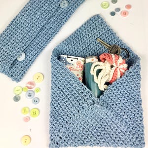 May include: A light blue crocheted envelope pouch with a zipper closure. The pouch is filled with various items, including a small bottle of hand sanitizer, a key chain, and a small crocheted toy. The pouch is sitting on a white surface.