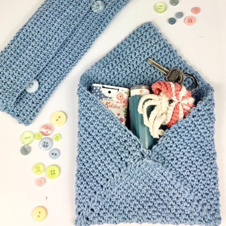 May include: A light blue crocheted envelope pouch with a zipper closure. The pouch is filled with various items, including a small bottle of hand sanitizer, a key chain, and a small crocheted toy. The pouch is sitting on a white surface.