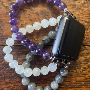 May include: Three beaded bracelets in amethyst, white, and gray colors, attached to a black Apple Watch.