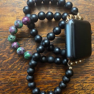 May include: A black beaded phone strap with a gold clasp and a black smartwatch attached. The strap is made of round beads and is wrapped around the smartwatch.