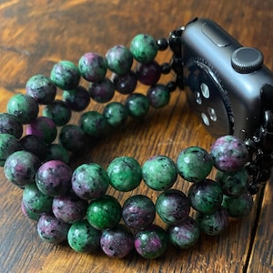 May include: A black Apple Watch band made of green and pink gemstone beads. The band is made of three rows of beads and has a black metal clasp.