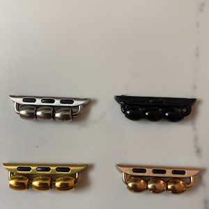 K&ouml;nnte beinhalten: Vier Apple Watch Band-Adapter in verschiedenen Ausf&uuml;hrungen: Silber, Schwarz, Gold und Ros&eacute;gold. Jeder Adapter hat drei runde Perlen und einen rechteckigen Einsatz mit vier schwarzen rechteckigen Ausschnitten.